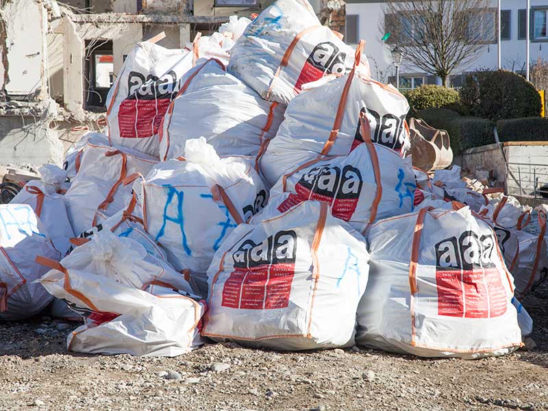 Mehrere Big Bags mit Asbest Warnung liegen auf einem Haufen vor einer Baustelle.