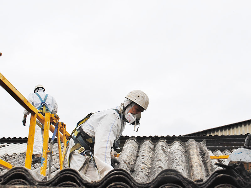 Zwei Personen mit Schutzkleidung arbeiten mit Asbest auf dem Dach.