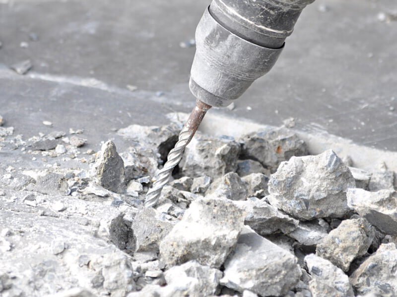 Staubbedeckte Bohrmaschine inmitten von Betonplatten sowie Betonabbruch.