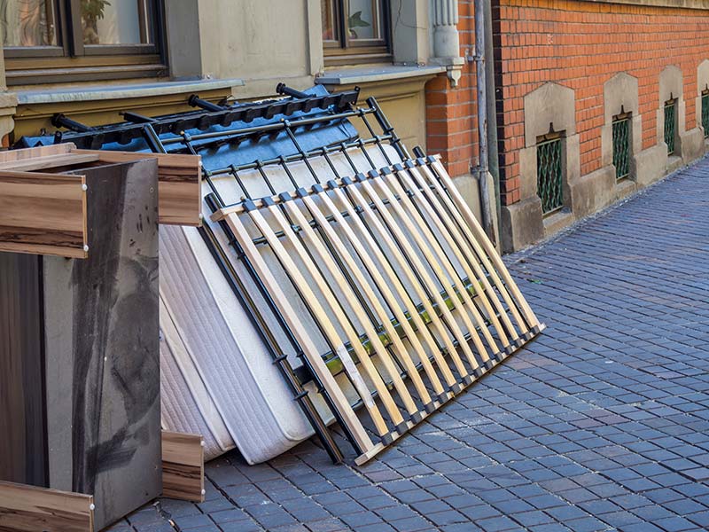 Ein Bett mit Lattenrost und Matratze lehnt an einer Hausfassade und wartet auf die Entsorgung.