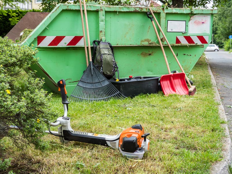 Grüner Container für Gartenabfälle steht auf einer Wiese neben Gartenwerkzeuge.