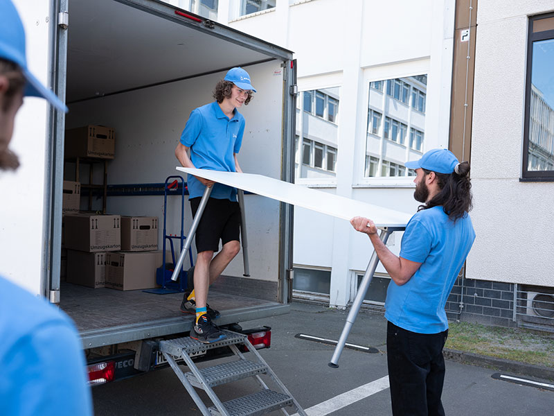 Zwei Personen tragen einen Tisch in einen bereits mit einigen Umzugskartons gefüllten Transporter.