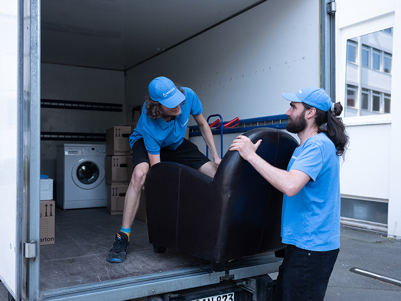 Zwei Personen tragen einen Sessel in einen mit verschiedenen Gegenständen befüllten Transporter.