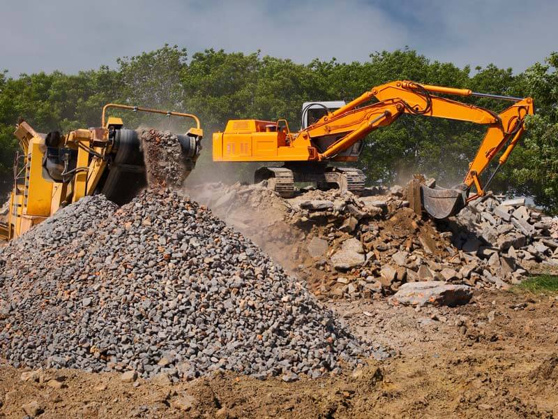 Ein Bagger führt Bauschutt in eine Aufbereitungsanlage, aus der zerkleinerte Steine auf einem Fließband heraustransportiert werden.