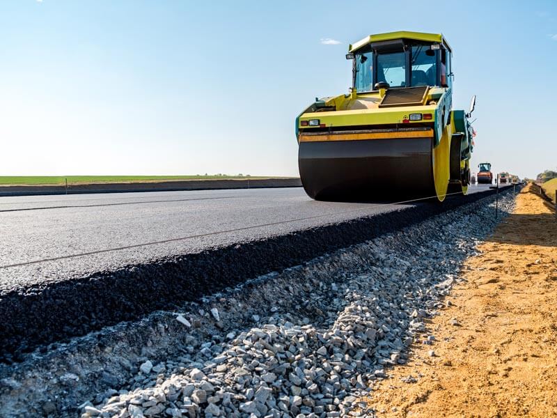 Eine Walze fährt auf einer frisch asphaltierten Straße. Neben dem Asphalt ist eine Unterschicht aus Schotter erkennbar.