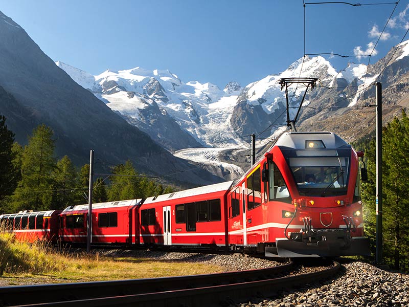 Ein Zug fährt durch eine Alpenlandschaft mit schneebedeckten Bergen im Hintergrund.