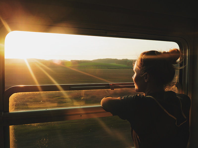 Person steht an einem geöffneten Fenster in einem Zug und schaut den Sonnenuntergang an.